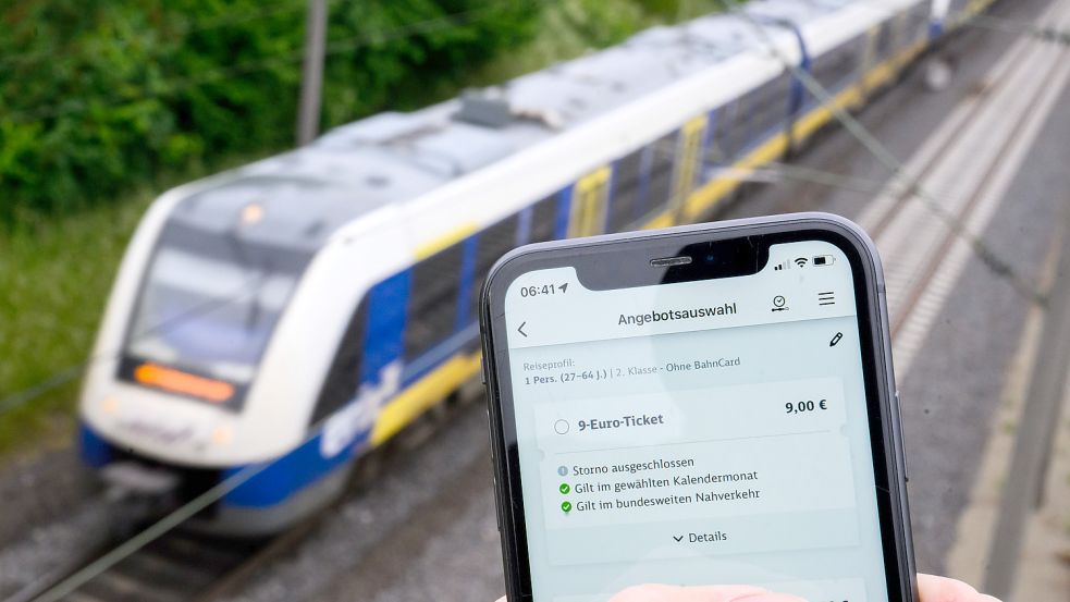 Auf einem Smartphonedisplay ist die Bahn-App „DB Navigator“ zu sehen, in der man das Neun-Euro-Ticket digital kaufen kann. Im Hintergrund fährt ein Regionalzug vorbei Foto: Stratenschulte/DPA