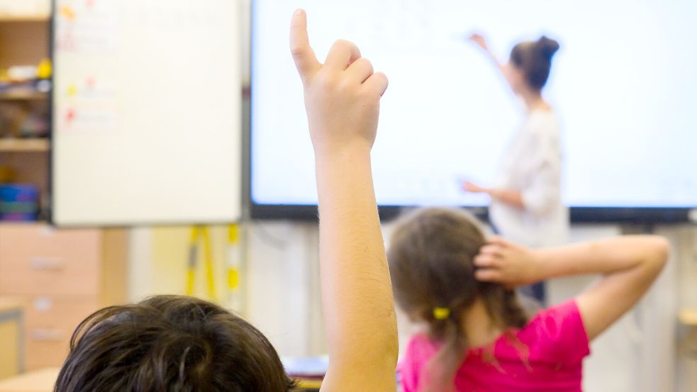 Ein Schüler meldet sich per Handzeichen während eine Lehrerin auf eine digitale Schultafel im Klassenraum einer 4. Klasse schreibt. Foto: Julian Stratenschulte/dpa